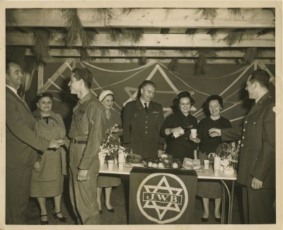 2001.040.017 – This photo from 1959 depicts members of the Ladies of the Holiday Committee of the Baltimore Jewish Welfare Board serving wine and cake in a sukkah to Jewish troops of the Aberdeen Proving Ground.