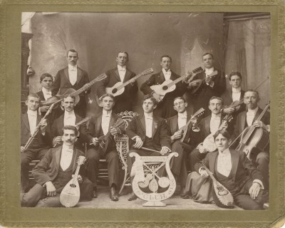 The Washburn Club, circa 1900.  Each member is holding an instrument, primarily strings; their logo, featured in the cardboard cut-out in the center front, consists of a mandolin, guitar, and banjo within a lyre.  Bonus: spot the disembodied hand holding on to the backdrop in the upper left.  Gift of Mr. and Mrs. Morton K. Sugar. JMM 1987.193.2