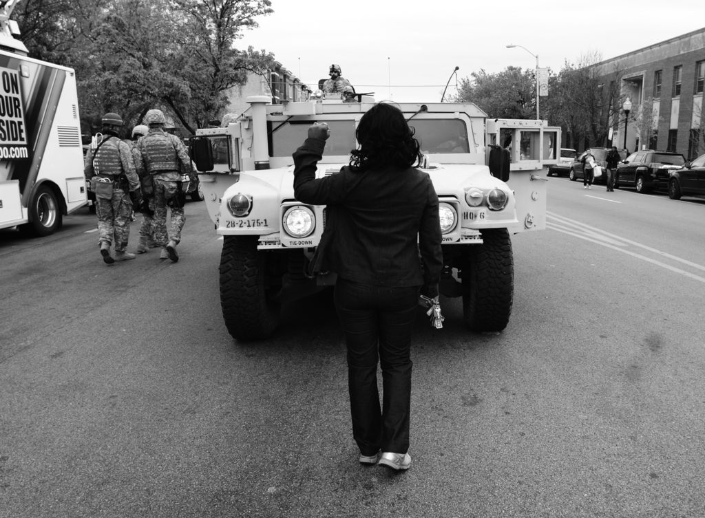 A black and white photo of a person standing in front of an armored truck, holding their fist up. They are turned away from the camera.