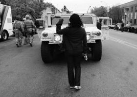 A black and white photo of a person standing in front of an armored truck, holding their fist up. They are turned away from the camera.