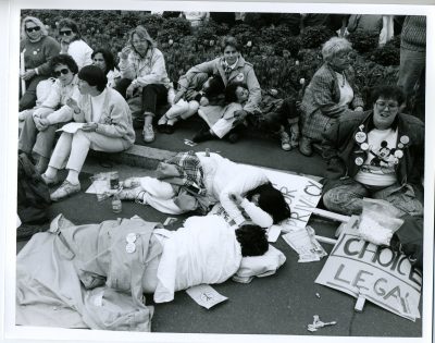 In this black and white image, a group of people sit near the road, on the curb. Two of the people in the image are laying down on the road, asleep. There is a sign that says "Choice. Legal"