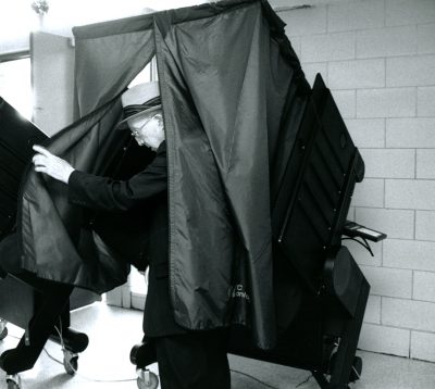In this black and white image, an older, white man steps out of a polling booth, which has curtains he's pulling aside for privacy.