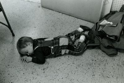 In this black and white photo, a toddler lays asleep on a floor, near a pile of coats.