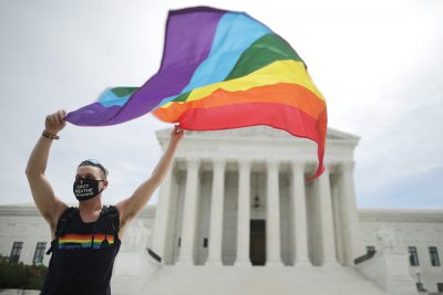 A white person stands outside the US Supreme Court building. They are wearing a mask and holding a rainbow Pride flag.