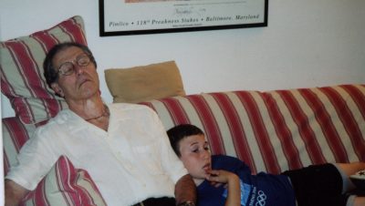 A man and a boy sit on a red and white striped couch. The boy is leaning against the man who has fallen asleep.