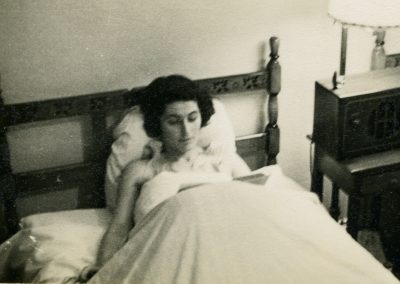 A woman sits in bed, reading a book. Her legs are covered by a blanket.et.