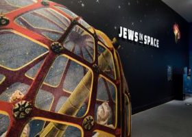 A photograph of the Jews in Space exhibit, showing a feature wall with a starscape and an illustration of people looking through a telescope. The words “Jews in Space” are in white on the star scape.