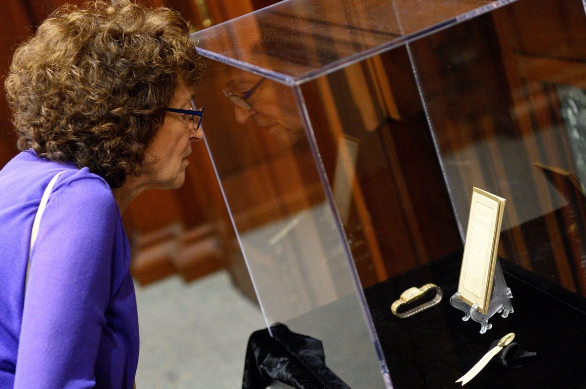 A person looks into a display case that holds a pamphlet, a watch, and a ribbon. The person leans in closely to look.
