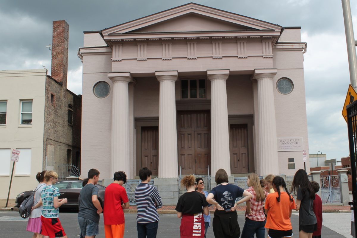 A group of young people stand outside the Lloyd Street Synagogue, a large building with columns and a pointed top. The group faces a tour guide who speaks to the group.