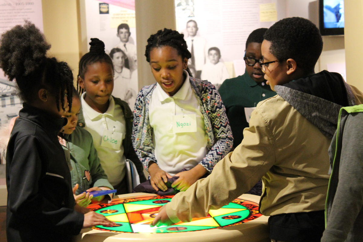 A group of young students stand in the basement of the Lloyd Street Synagogue, working on the stained glass window hands-on activity together.