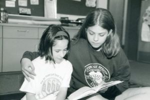 A black and white image of two girls sitting together reading a book. One of the girls has her arm around the other and they look down at the book in her lap together.