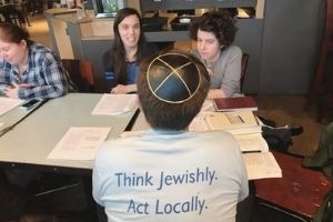 A group of white people sit at a table, talking together. THere are books and papers on the table. One person, in the foreground, is facing away from the camera, and they are wearing a yarmulke and a shirt that says “Think Jewishly. Act Locally.” on the back.