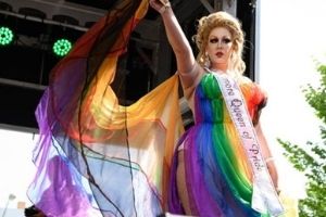 A photograph of Drag Queen Bambi Galore. She is dressed in a flowing, rainbow dress that she flounces while on stage. She also wears a sash that says “Baltimore Queen of Pride”.