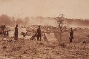 A sepia-tone photo of soldiers standing outside a civil war encampment, dressed in uniform. In the distance there are more tents and a tree-line.