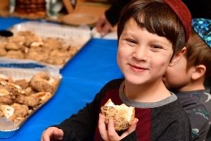 A young, white boy, looks at the camera smiling. He holds a baked good that he has taken bites out of and wears a yarmulke. There are more baked goods on baking sheets in the background, on a table.