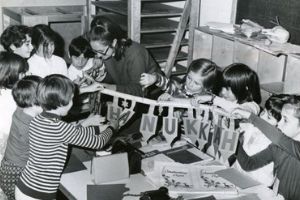A black and white photo of a group of children who are working on an art project together, sitting around a table with their teacher. They hold up a banner that says Hanukkah on it.