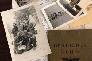 A photo of a pile of papers, including photographs. The photograph in focus shows a group of men standing together, wearing uniforms. There is also a folder that says “Deutches Reich” next to the photograph.