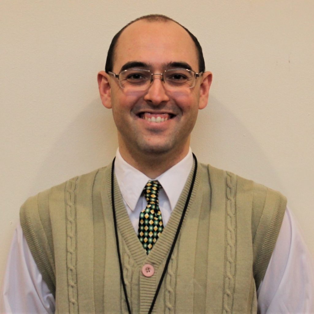 A white man with short, dark hair, glasses, wearing a button-up shirt, tie, and sweater vest. He stands in front of a white background. He is smiling.
