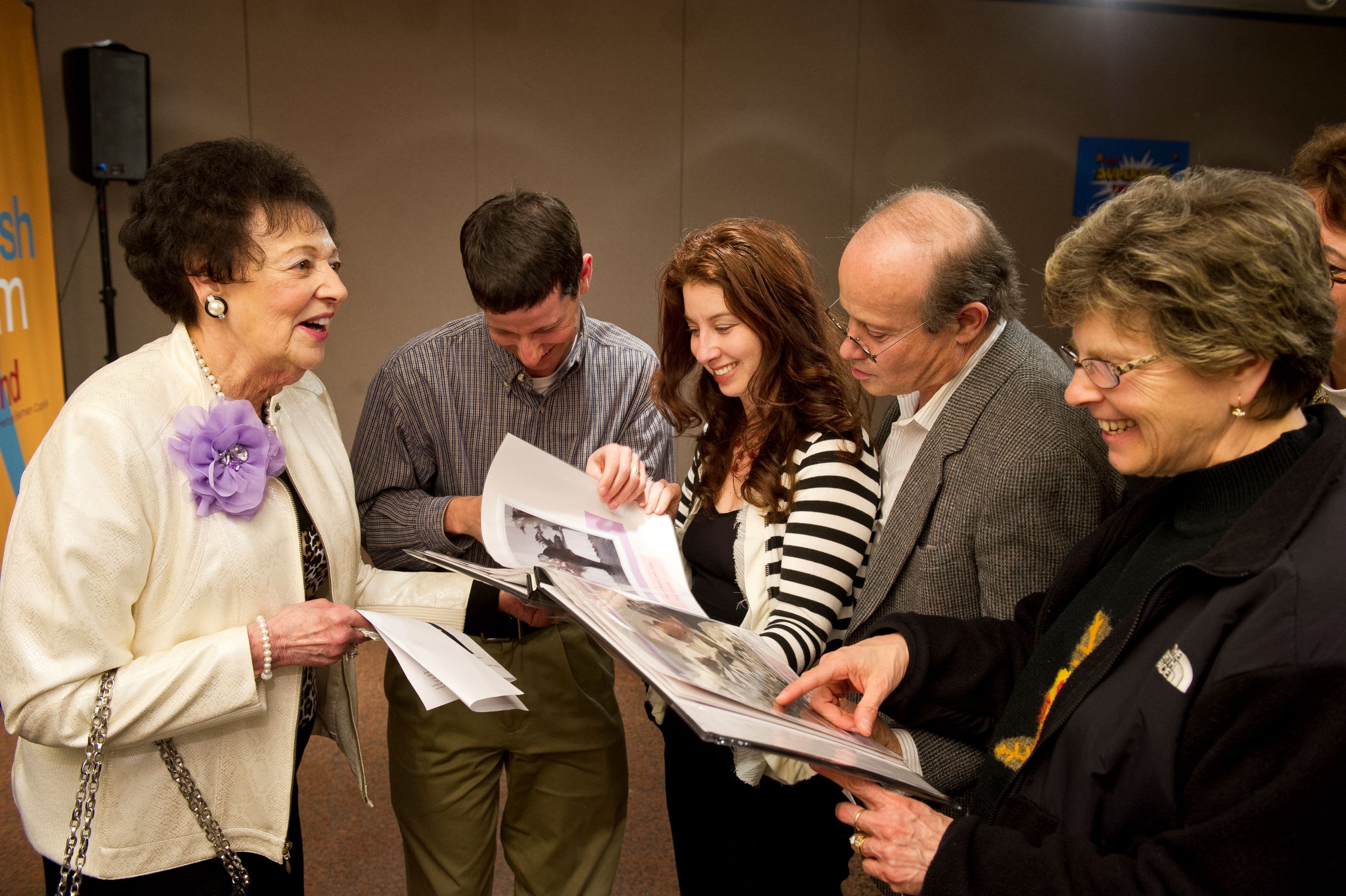 A group of people stand together talking and smiling. They look at a book filled with images. One person points to an image.
