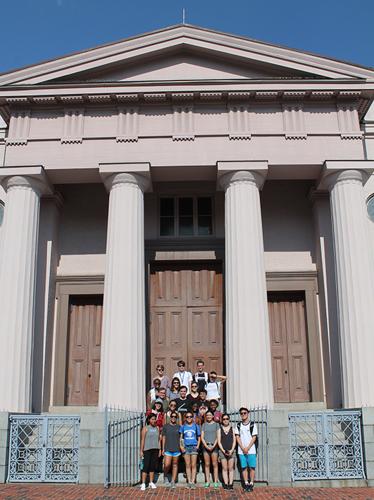 A group of visitors stand on the exterior steps of the Lloyd Street Synagogue. The synagogue is a large building with columns and a pointed top.