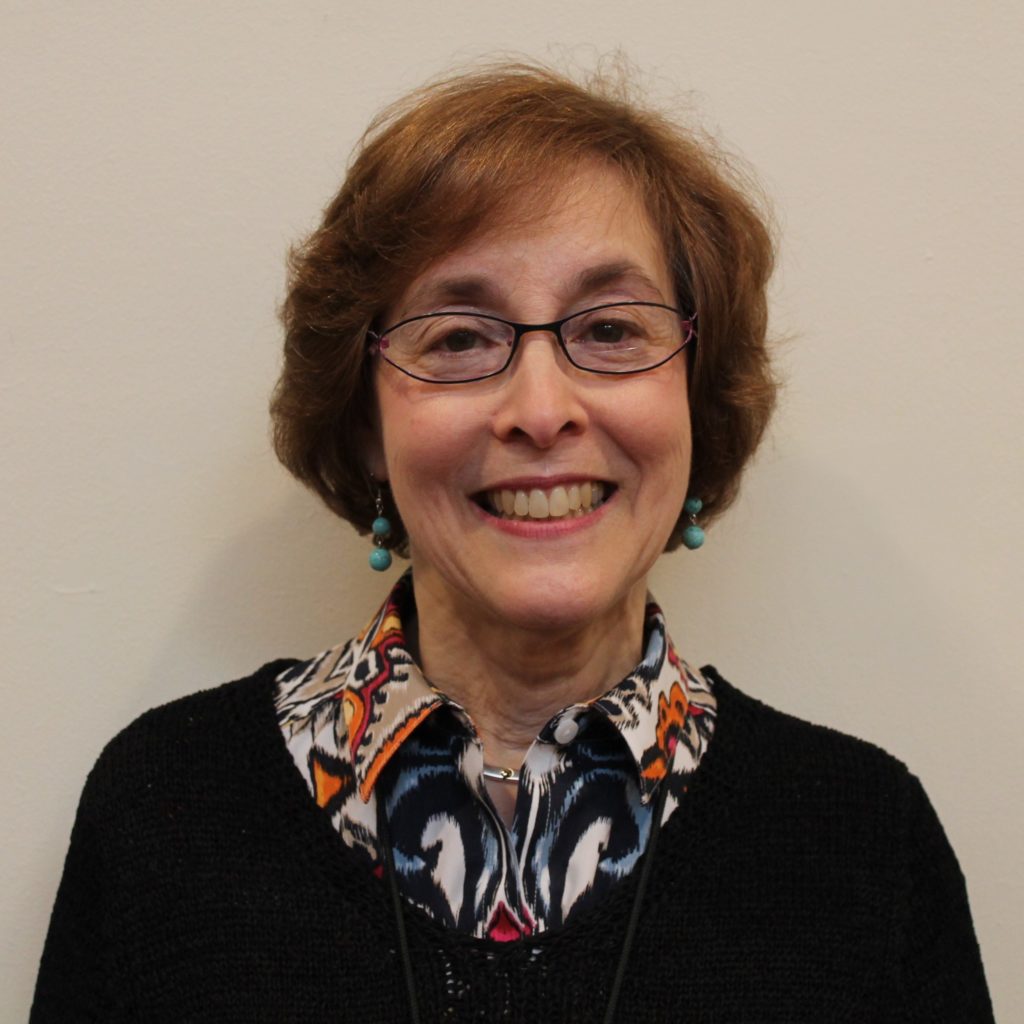A white woman with short, brown hair, glasses, wearing a black sweater and a patterned blouse. She stands in front of a white background. She is smiling.