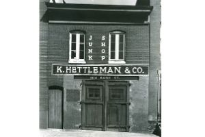 A black and white photo of a short, two-story building that has a sign that says “K. Helltman & Co.” on it, hanging over the double front doors.