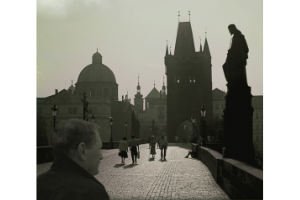 A black and white photo of a bridge in Prague, showcasing the architecture of the city. A white man stands in the foreground, his back towards the camera, looking across the bridge.
