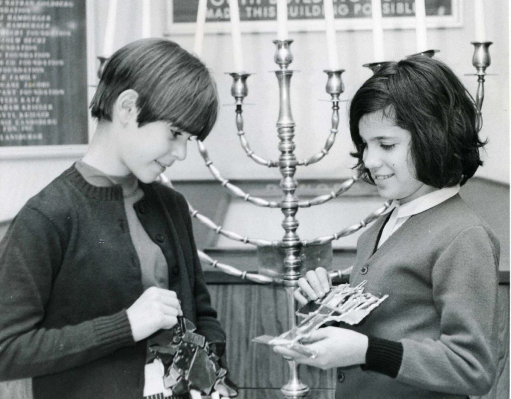 Two white girls stand in front of a large menorah. They are both holding Hanukkah related handicrafts and look like they are showing them to each other.