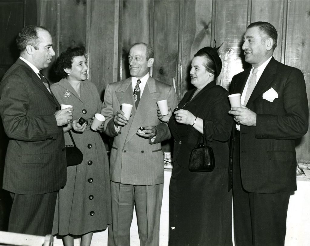 Black and white photo of a group of 5 white adults, three men and two women stand holding paper drink cups. Some are holding what look like desserts in their hands. All are smiling at each other.