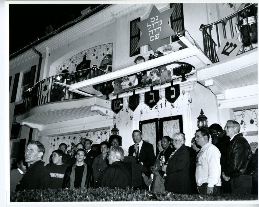 black and white photograph of Mayor Martin O'Malley standing at a podium in front of the doors to a house decorated with all sorts of Hanukkah themed items. He is surrounded by a small grouwd listening to him speak.