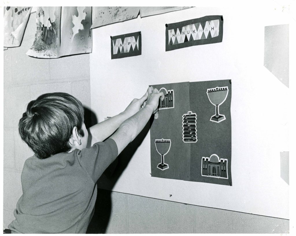 Black and white photo of a young white boy sticking some Hanukkah decorations on a wall. He is facing away from the camera.