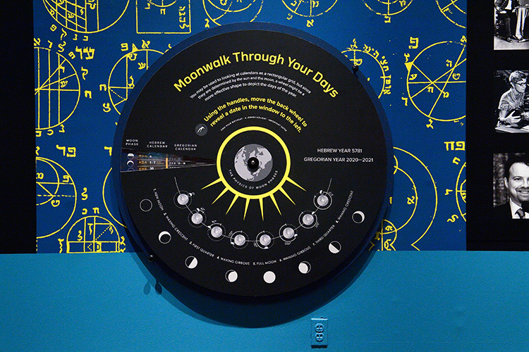 A circular hands-on activity in our Jews in Space exhibit. The words “Moonwalk Through Your Days” are in yellow at the top of the wheel. There are knobs you can spin to change the dates in the window on the wheel.