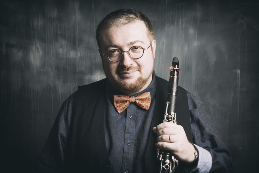 A white man wearing glasses and an orange bowtie holds a clarinet.