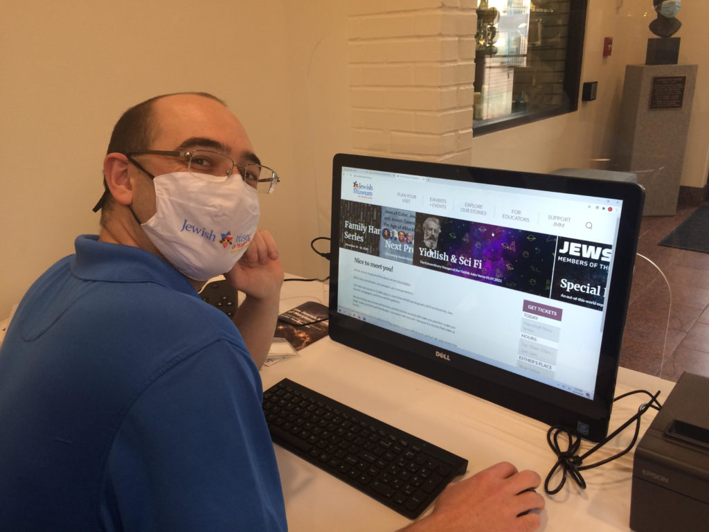 color photo of a young white man wearing classes and a white mask printed with the Jewish Museum of Maryland logo. He is seated in front of a large computer screen which shows the homepage of the JMM website.