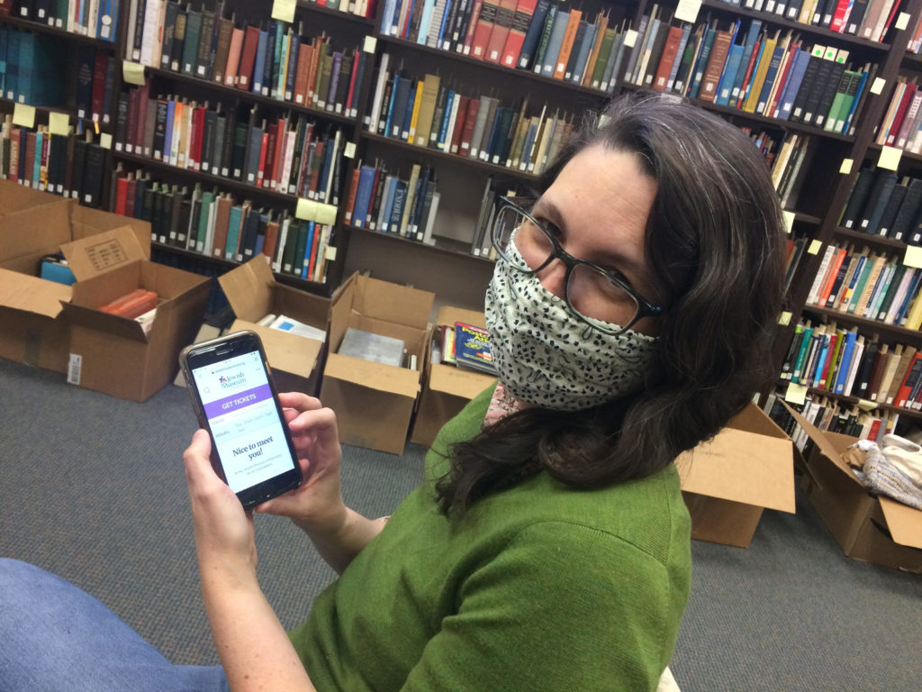 Color photo of a white woman with dark hair wearing a polka dot printed mask and glasses. She is holding a smart phone with the JMM website mobile version homescreen showing.