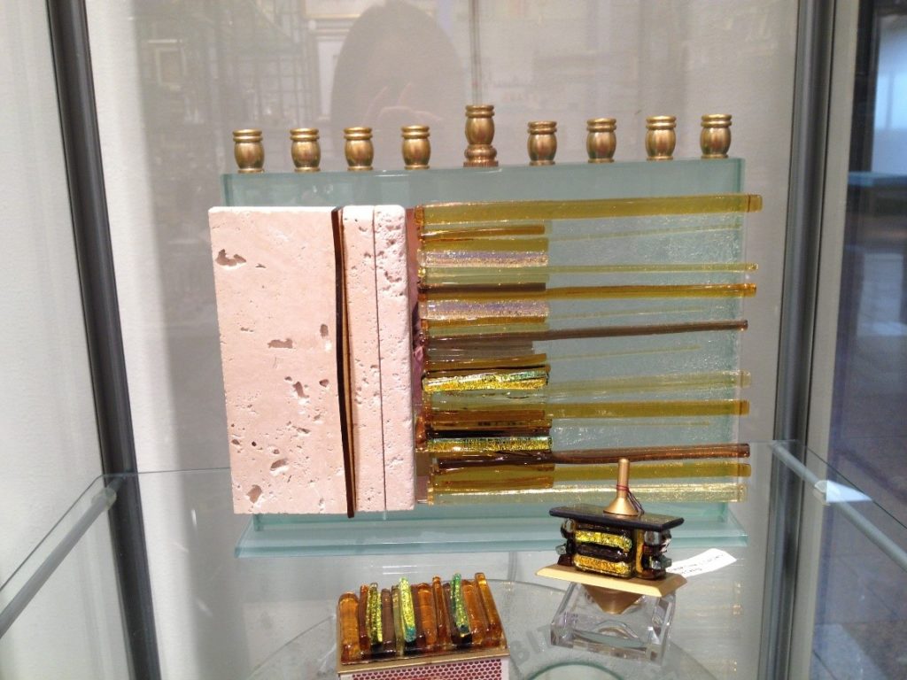 Color photograph showing a menorah, dreidel, and matchbox, all in the same style combining colored glass and stone.