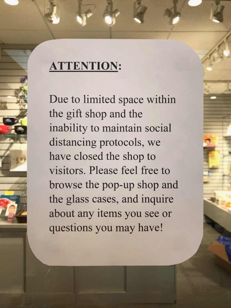 A white paper sign with rounded counters hung up on a glass door. Sign reads "Attention: Due to limited space within the gift shop and the inability to maintain social distancing protocols, we have closed the shop to visitors. Please feel free to browse the pop-up shop and the glass cases, and inquite about any items you see or questions you may have!)