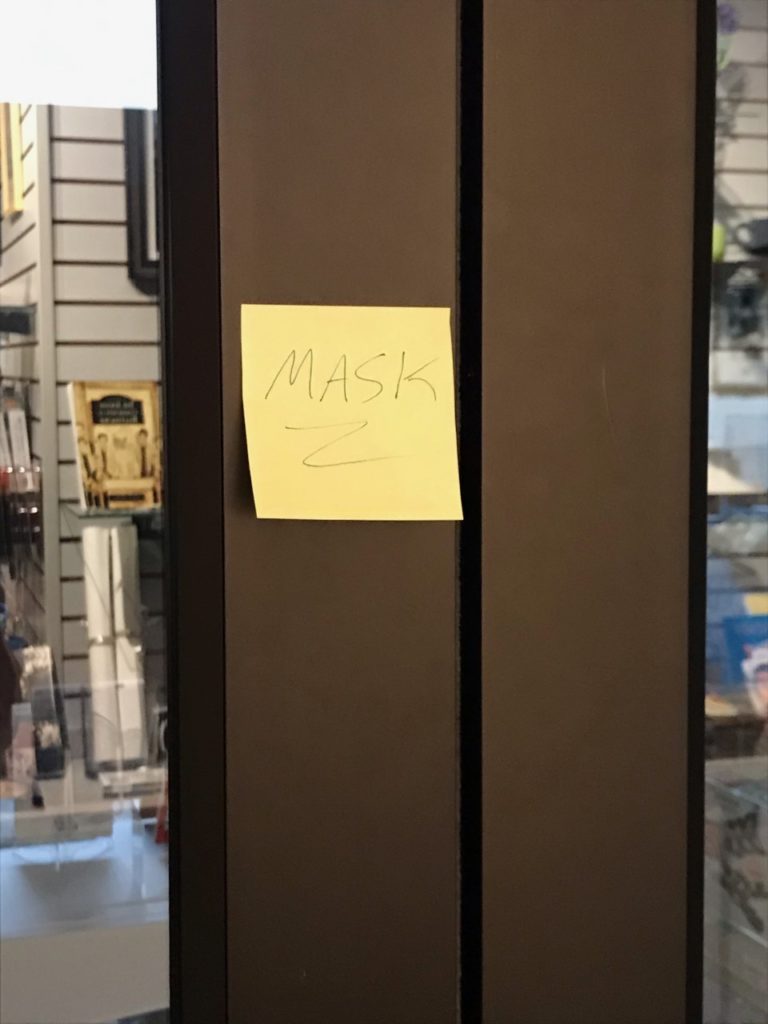 A small yellow sticky note reading "Mask" placed on the frame of a double doorway.
