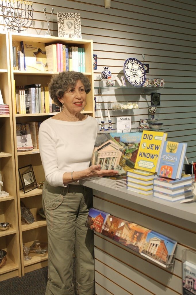 An older white woman with short curly hair stands next to a shop counter pointing to a display of books, including a children's book about the Lloyd Street Synagogue, "Did Jew Know," and a book on Jewish holidays