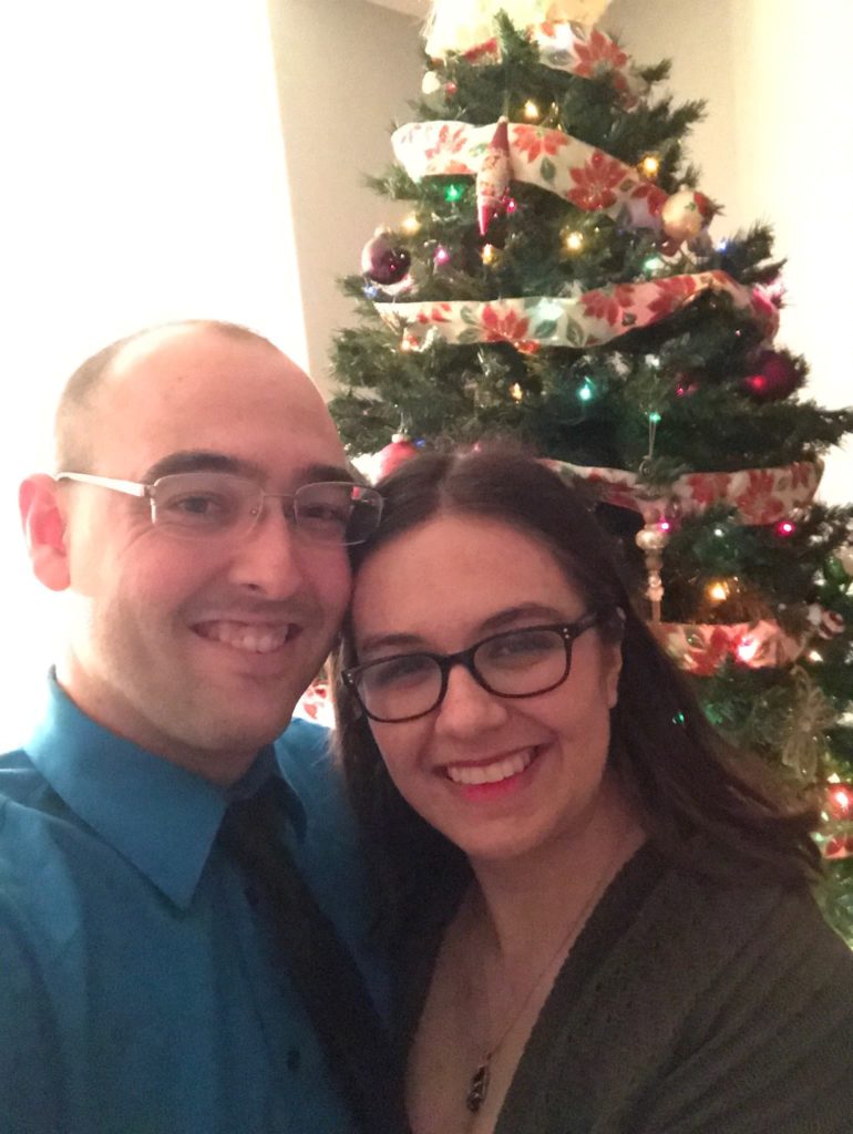 A young white couple, man on the left, woman on the right, both wearing glasses, stand in front of a decorated Christmas tree. They are both smiling at the camera.