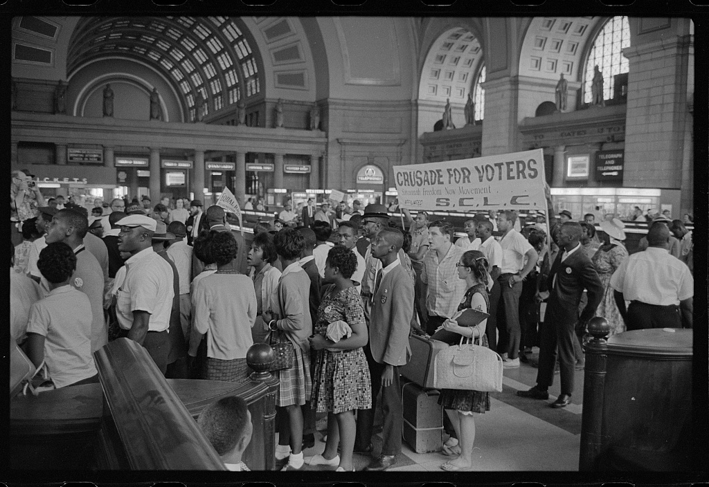 Black and white photo showing a large crowd of mostly Black people standing inside a building with tall, arched ceilings. A side is being held up that reads “Crusade for voters Savannah Freedom Now Movement S.C.L.C.”