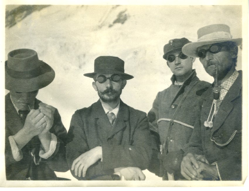 Four unidentified individuals facing the camera and standing in the snow, all four are wearing heavy coats and goggles.