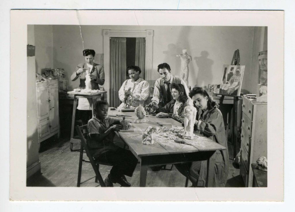 5 students—two Black women, two women of indeterminate race, and one Black boy—create sculptures in a workroom. In the middle of the photo, teaching artist Reuben Kramer, a white man, looks toward the sculpture being made by the young boy.