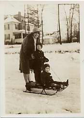 Two children on a toboggan-like sled with an adult pushing them forward.