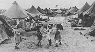 A group of children stand between rows of tents, which have clotheslines hanging between them.