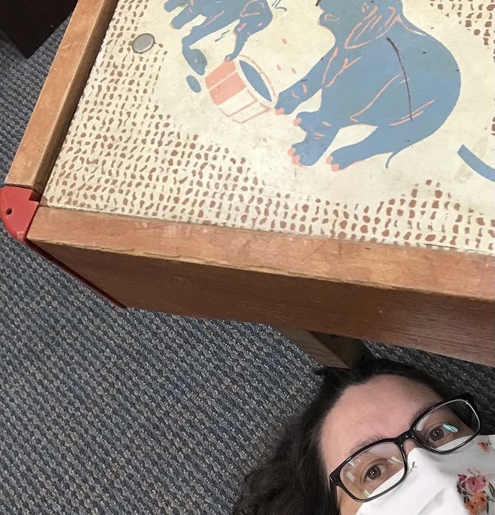 A woman in glasses and a floral mask lies on the floor next to a piece of vintage baby furniture, with an elephant on it.