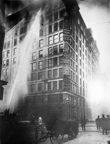 Black and white image of a large building on fire being sprayed with fire hoses. 