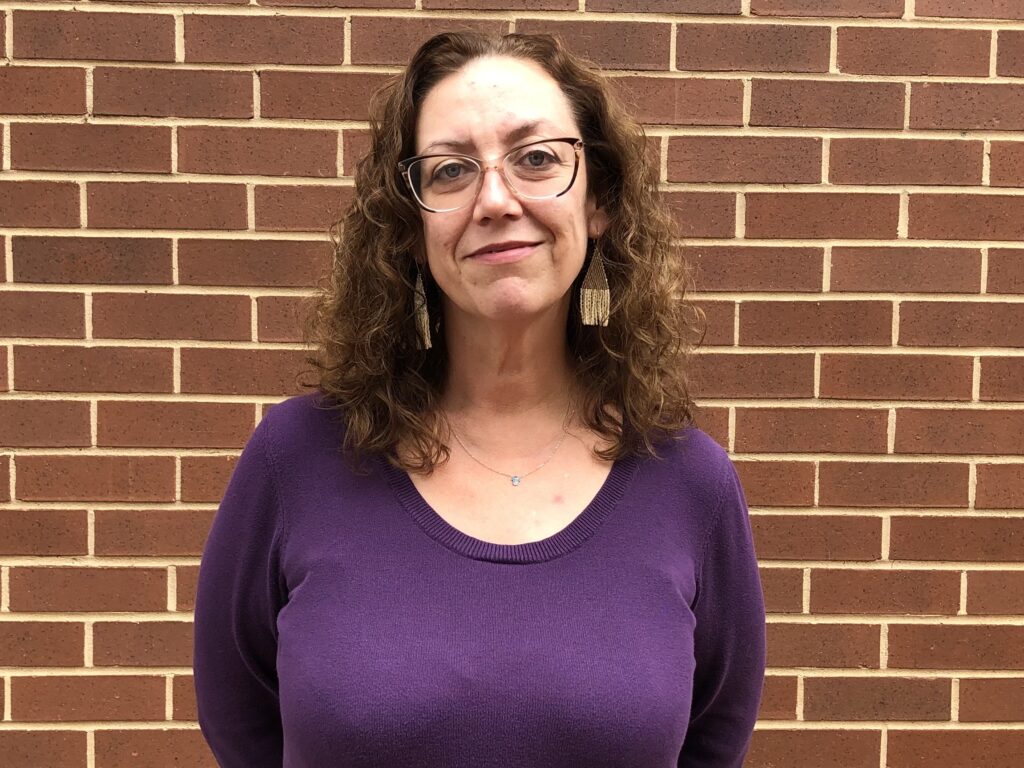 A white woman wearing glasses, earrings, and a purple shirt stands in front of a brick wall.
