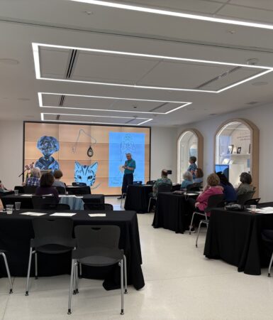 People sit at tables facing a large screen, where a man stands holding a microphone