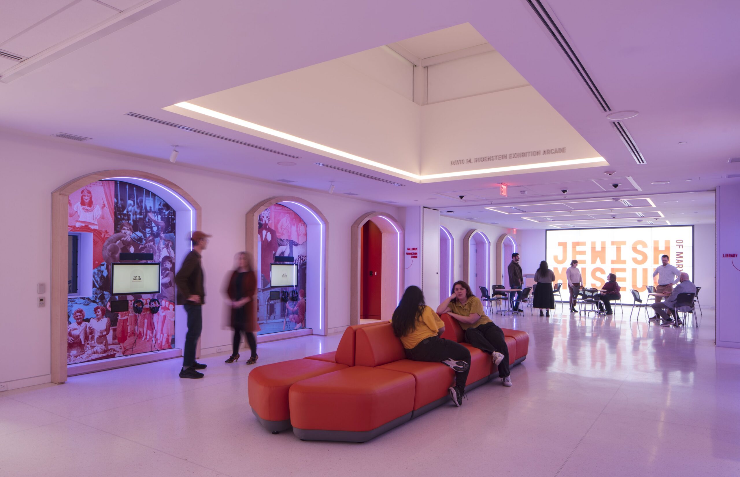 Image of the interior of a Museum. The room is lit with a purple tone and a large seating area occupies the center of the floor. Several people are in frame, in conversation with one another and moving through the space.